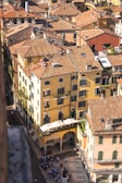 An aerial view of a historical European cityscape with tightly packed buildings featuring terracotta roofs. A yellow building with arched windows and a terrace with white umbrellas is prominently seen in the foreground. A cobblestone street runs alongside the building, where a group of people gathers.