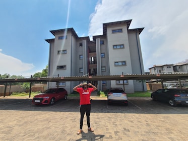 A multi-story residential building with a modern design stands in the background, featuring large windows and beige walls. In the foreground, a person in a red shirt is standing with their hands on their head. Several cars are parked under a carport, including a red and a blue car. The scene is set under a bright, clear sky with some clouds.
