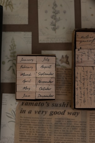 A collection of wooden stamps displaying the months of the year is arranged on a surface with botanical drawings and botanical-themed paper. The stamps are neatly aligned in two columns, showing months from January to December in a handwritten font. The background features images of plants and text from what appears to be a newspaper or magazine article.