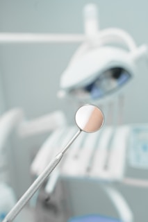 A dental mirror held by a textured metal handle is placed in focus with a blurred background displaying dental equipment. The setting appears to be a dental office or clinic, with soft lighting and a clean, sterile environment.