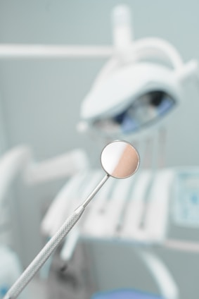 A dental mirror held by a textured metal handle is placed in focus with a blurred background displaying dental equipment. The setting appears to be a dental office or clinic, with soft lighting and a clean, sterile environment.