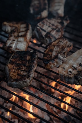 Several pieces of grilled meat are cooking on a barbecue grill over glowing hot coals. The meat appears to be partially charred with visible grill marks.