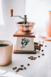 An old wooden coffee grinder resting on a traditional woven mat beside a steaming cup of drip coffee.