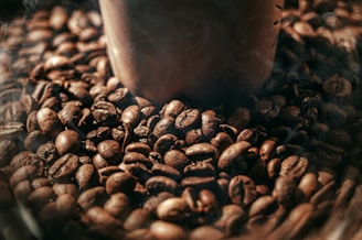 A close-up view of a pile of roasted coffee beans surrounding the base of a brown coffee cup. The beans have a shiny surface and rich, dark brown color, suggesting freshness and a roasted aroma.