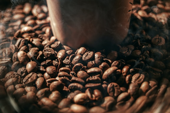 A close-up view of a pile of roasted coffee beans surrounding the base of a brown coffee cup. The beans have a shiny surface and rich, dark brown color, suggesting freshness and a roasted aroma.
