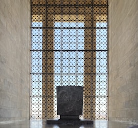A large, ornate metal screen with a floral pattern allows light to pass through, casting shadows on a tall stone structure in front. The room is expansive, with high ceilings and walls made of light-colored stone.