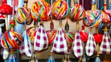A collection of colorful fabrics and yarns arranged neatly on wooden sticks. The fabrics feature vibrant, multicolored stripes and check patterns, predominantly in reds, blues, yellows, and greens. The yarns are wrapped tightly around the sticks, creating spherical shapes.