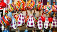 A collection of colorful fabrics and yarns arranged neatly on wooden sticks. The fabrics feature vibrant, multicolored stripes and check patterns, predominantly in reds, blues, yellows, and greens. The yarns are wrapped tightly around the sticks, creating spherical shapes.