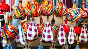 A collection of colorful fabrics and yarns arranged neatly on wooden sticks. The fabrics feature vibrant, multicolored stripes and check patterns, predominantly in reds, blues, yellows, and greens. The yarns are wrapped tightly around the sticks, creating spherical shapes.
