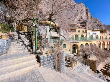 Stone steps lead up to a terrace area with potted plants and bare trees, set against a backdrop of rocky cliffs. The architecture features arched windows and doors, with green railings and a mix of stone and brick materials.