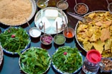 An assortment of food items is displayed on a table, featuring a variety of spices in small bowls, fresh greens on plates, and crunchy bread pieces arranged on a large tray. There are also bowls filled with creamy white cheese-like substance, a mix of powdered spices, dried rose petals, and raisins. A transparent jug filled with a dark liquid is placed on the side.