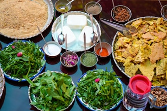 An assortment of food items is displayed on a table, featuring a variety of spices in small bowls, fresh greens on plates, and crunchy bread pieces arranged on a large tray. There are also bowls filled with creamy white cheese-like substance, a mix of powdered spices, dried rose petals, and raisins. A transparent jug filled with a dark liquid is placed on the side.
