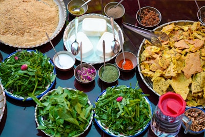 An assortment of food items is displayed on a table, featuring a variety of spices in small bowls, fresh greens on plates, and crunchy bread pieces arranged on a large tray. There are also bowls filled with creamy white cheese-like substance, a mix of powdered spices, dried rose petals, and raisins. A transparent jug filled with a dark liquid is placed on the side.