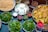 An assortment of food items is displayed on a table, featuring a variety of spices in small bowls, fresh greens on plates, and crunchy bread pieces arranged on a large tray. There are also bowls filled with creamy white cheese-like substance, a mix of powdered spices, dried rose petals, and raisins. A transparent jug filled with a dark liquid is placed on the side.