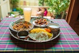 A vibrant and colorful meal is arranged on a tray atop a checkered tablecloth. The spread includes two dishes with rich curries, a portion of yellow and white rice, a side of yogurt, and a large glass of a pale drink. The setting appears to be outdoors, with blurred greenery and people visible in the background, adding a lively and inviting atmosphere.