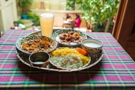 A vibrant and colorful meal is arranged on a tray atop a checkered tablecloth. The spread includes two dishes with rich curries, a portion of yellow and white rice, a side of yogurt, and a large glass of a pale drink. The setting appears to be outdoors, with blurred greenery and people visible in the background, adding a lively and inviting atmosphere.