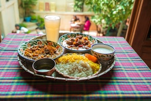 A vibrant and colorful meal is arranged on a tray atop a checkered tablecloth. The spread includes two dishes with rich curries, a portion of yellow and white rice, a side of yogurt, and a large glass of a pale drink. The setting appears to be outdoors, with blurred greenery and people visible in the background, adding a lively and inviting atmosphere.