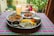 A vibrant and colorful meal is arranged on a tray atop a checkered tablecloth. The spread includes two dishes with rich curries, a portion of yellow and white rice, a side of yogurt, and a large glass of a pale drink. The setting appears to be outdoors, with blurred greenery and people visible in the background, adding a lively and inviting atmosphere.