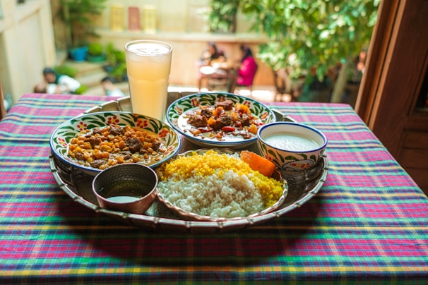 A vibrant and colorful meal is arranged on a tray atop a checkered tablecloth. The spread includes two dishes with rich curries, a portion of yellow and white rice, a side of yogurt, and a large glass of a pale drink. The setting appears to be outdoors, with blurred greenery and people visible in the background, adding a lively and inviting atmosphere.