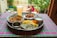 A vibrant and colorful meal is arranged on a tray atop a checkered tablecloth. The spread includes two dishes with rich curries, a portion of yellow and white rice, a side of yogurt, and a large glass of a pale drink. The setting appears to be outdoors, with blurred greenery and people visible in the background, adding a lively and inviting atmosphere.