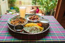 A vibrant and colorful meal is arranged on a tray atop a checkered tablecloth. The spread includes two dishes with rich curries, a portion of yellow and white rice, a side of yogurt, and a large glass of a pale drink. The setting appears to be outdoors, with blurred greenery and people visible in the background, adding a lively and inviting atmosphere.
