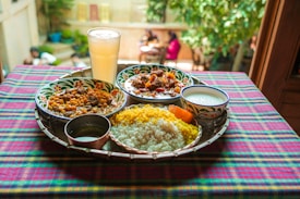 A vibrant and colorful meal is arranged on a tray atop a checkered tablecloth. The spread includes two dishes with rich curries, a portion of yellow and white rice, a side of yogurt, and a large glass of a pale drink. The setting appears to be outdoors, with blurred greenery and people visible in the background, adding a lively and inviting atmosphere.