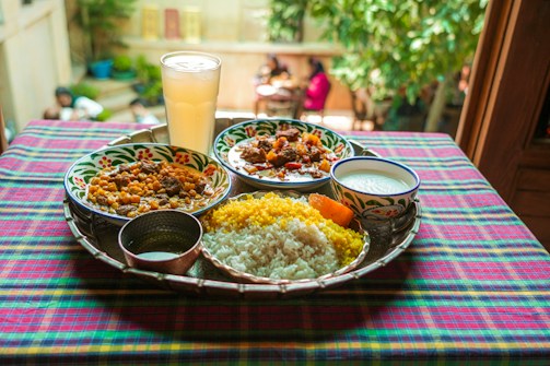 A vibrant and colorful meal is arranged on a tray atop a checkered tablecloth. The spread includes two dishes with rich curries, a portion of yellow and white rice, a side of yogurt, and a large glass of a pale drink. The setting appears to be outdoors, with blurred greenery and people visible in the background, adding a lively and inviting atmosphere.