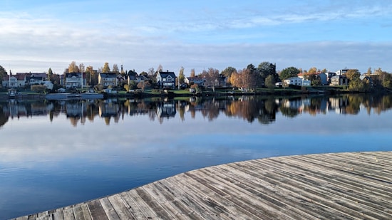 A serene lakeside view with a row of houses along the shoreline. The water reflects the houses and surrounding trees, which display autumnal colors. A wooden deck in the foreground curves gently, adding depth to the scene. The sky is partly cloudy, casting a soft light over the landscape.