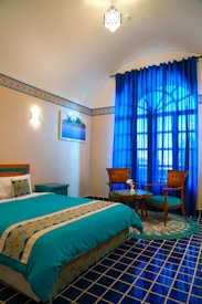A well-decorated bedroom featuring a turquoise-themed bedspread and matching pillows, with a painting hanging above a small nightstand. Two wooden chairs with turquoise cushions and a small round table sit beside a large window draped with bright blue curtains. The room has a patterned blue and white tiled floor and decorative wall tiles that add to the aesthetic appeal.