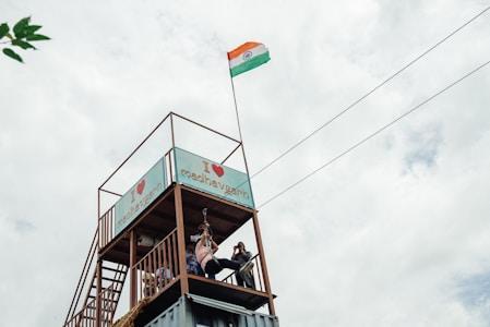 A raised platform with a structure that has a sign reading 'I ❤️ madhavgarh'. A person is zip-lining, and an Indian flag is prominently displayed on a tall pole, waving in the wind. The sky is overcast with clouds, creating a grayish backdrop.
