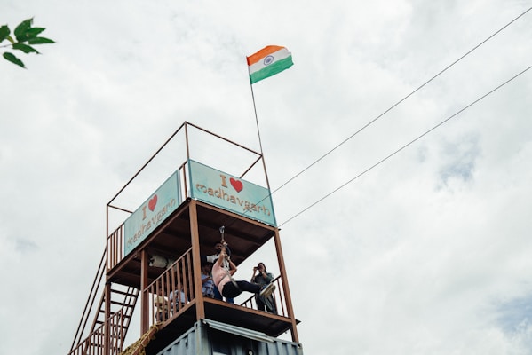 A raised platform with a structure that has a sign reading 'I ❤️ madhavgarh'. A person is zip-lining, and an Indian flag is prominently displayed on a tall pole, waving in the wind. The sky is overcast with clouds, creating a grayish backdrop.