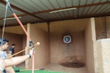 An indoor archery range with a person holding a drawn bow aimed at a colorful target positioned against a stone wall. Another person stands in the background near the entrance. Arrows are visible on the ground in front of the target.
