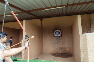 An indoor archery range with a person holding a drawn bow aimed at a colorful target positioned against a stone wall. Another person stands in the background near the entrance. Arrows are visible on the ground in front of the target.