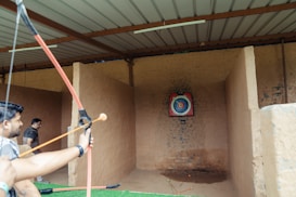 An indoor archery range with a person holding a drawn bow aimed at a colorful target positioned against a stone wall. Another person stands in the background near the entrance. Arrows are visible on the ground in front of the target.