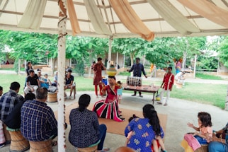 A vibrant outdoor performance is taking place under a large tent. A performer dressed in colorful, traditional attire is at the center, captured mid-dance, while a group of musicians accompanies them with drums. The audience, sitting on woven stools, is engaged and intently watching the performance. Lush green trees surround the area, adding a lively backdrop to the setting.