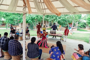 A vibrant outdoor performance is taking place under a large tent. A performer dressed in colorful, traditional attire is at the center, captured mid-dance, while a group of musicians accompanies them with drums. The audience, sitting on woven stools, is engaged and intently watching the performance. Lush green trees surround the area, adding a lively backdrop to the setting.