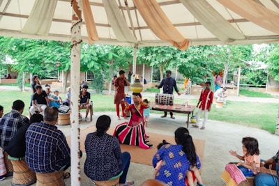 A vibrant outdoor performance is taking place under a large tent. A performer dressed in colorful, traditional attire is at the center, captured mid-dance, while a group of musicians accompanies them with drums. The audience, sitting on woven stools, is engaged and intently watching the performance. Lush green trees surround the area, adding a lively backdrop to the setting.