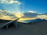 Rows of solar panels are aligned in a vast, arid landscape. The sun is setting or rising behind clouds, casting rays of light across the sky, while mountains can be seen in the distance.