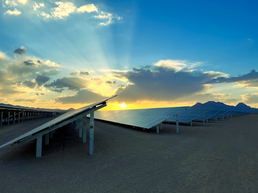 Rows of solar panels are aligned in a vast, arid landscape. The sun is setting or rising behind clouds, casting rays of light across the sky, while mountains can be seen in the distance.
