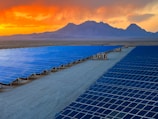 A vast array of solar panels stretches across a desert landscape under a vibrant orange and yellow sunset. The panels are arranged in neat rows, reflecting the sky's colors, and the background features distant mountains providing a sense of scale and isolation.