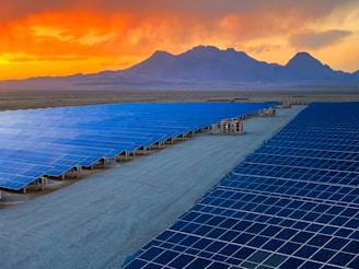 A vast array of solar panels stretches across a desert landscape under a vibrant orange and yellow sunset. The panels are arranged in neat rows, reflecting the sky's colors, and the background features distant mountains providing a sense of scale and isolation.
