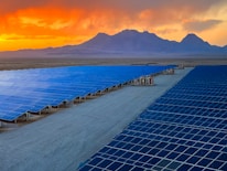 A vast array of solar panels stretches across a desert landscape under a vibrant orange and yellow sunset. The panels are arranged in neat rows, reflecting the sky's colors, and the background features distant mountains providing a sense of scale and isolation.