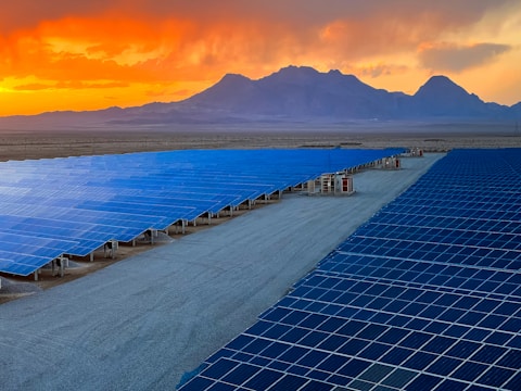 A vast array of solar panels stretches across a desert landscape under a vibrant orange and yellow sunset. The panels are arranged in neat rows, reflecting the sky's colors, and the background features distant mountains providing a sense of scale and isolation.