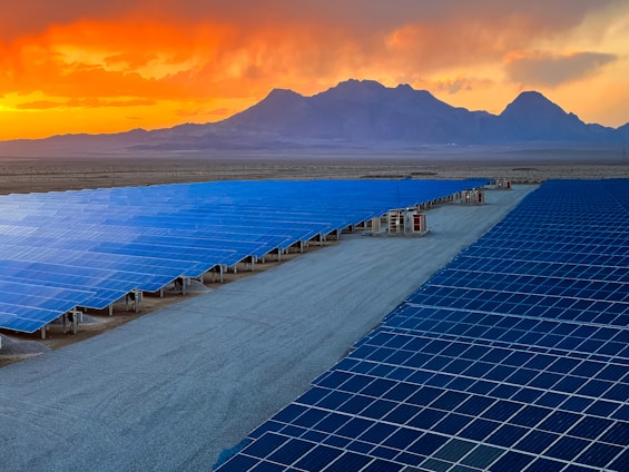 A vast array of solar panels stretches across a desert landscape under a vibrant orange and yellow sunset. The panels are arranged in neat rows, reflecting the sky's colors, and the background features distant mountains providing a sense of scale and isolation.