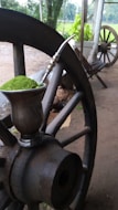 A metal mate cup filled with green yerba mate is placed on a large wooden wagon wheel. In the background, there are plants, a wooden bench, and another wheel, set in an outdoor rural environment.