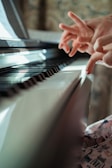 Hands are poised above a piano keyboard, with fingers lightly touching the keys. The lighting is warm and soft, casting a gentle glow across the instrument. The background shows a blurred pattern, adding depth to the focus on the hands and the piano.