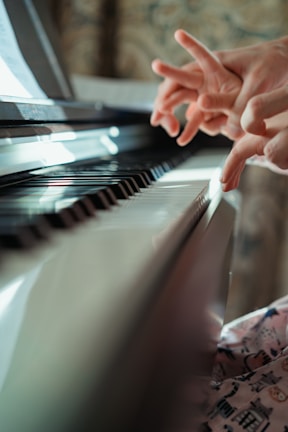 Hands are poised above a piano keyboard, with fingers lightly touching the keys. The lighting is warm and soft, casting a gentle glow across the instrument. The background shows a blurred pattern, adding depth to the focus on the hands and the piano.