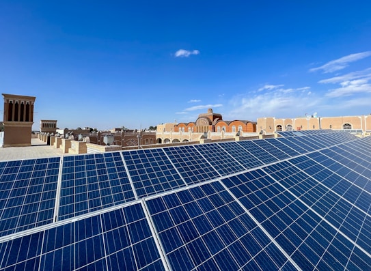 Large solar panels are prominently displayed on a rooftop, with traditional Middle Eastern architecture in the background under a clear blue sky. The setting includes a mix of modern technology and historical buildings.