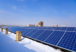 A row of solar panels is positioned on a rooftop, with snow partially covering the ground. The sky above is clear and blue, and several buildings are visible in the background.