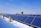 A row of solar panels is positioned on a rooftop, with snow partially covering the ground. The sky above is clear and blue, and several buildings are visible in the background.